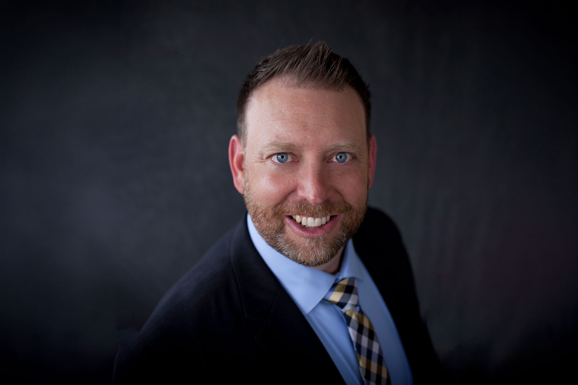 Shane Steadman smiling professionally in a dark suit with a white shirt and plaid tie against a dark background as a speaker for Apex Seminars.